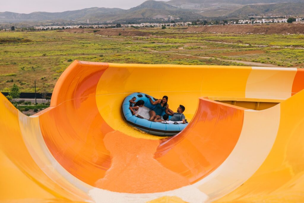 在野河尔湾尽情嬉戏4 big orange waterslide with blue inflatable sliding down with a family of four inside with vacant green lot and hills in the background