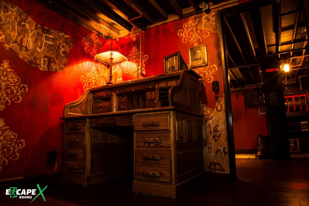 EscapeX Rooms 1 a room with a wooden desk with four drawers on each side and a space to place a chair with a red lighted lamp on top, with red wallpaper with golden artwork at escapex