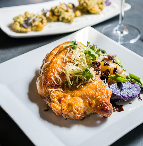 a square plate of chicken with garnish and a rectangular plate of vegetables and a glass of white wine with a vase filled with flowers at Andrei's Restaurant