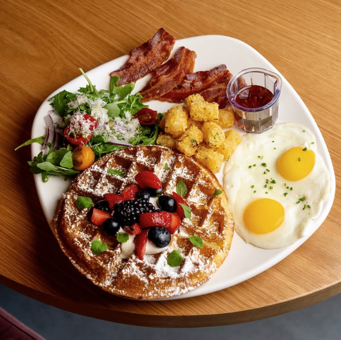 white plate with waffles with strawberries and berries on top and bacon, tater tots, two sunny side up eggs and a small portion of salad and syrup