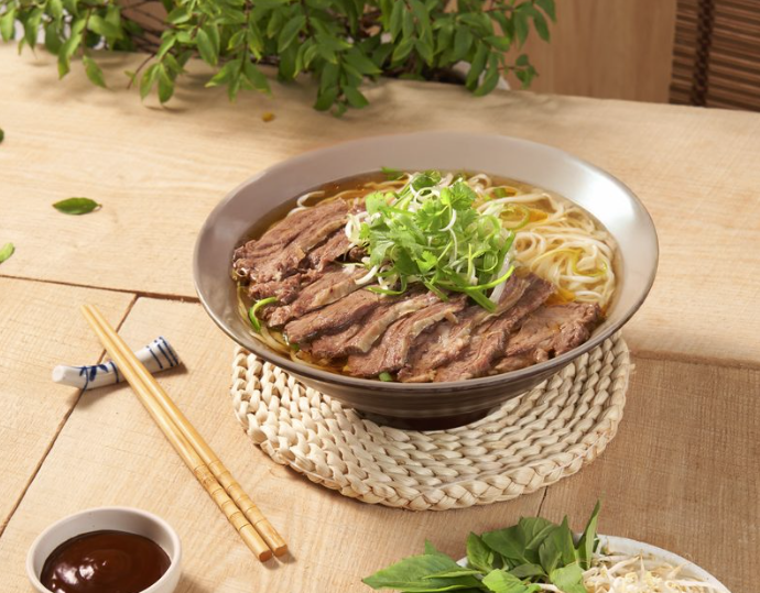 bowl of noodle soup with beef and green garnish on top, with two brown and red colored sauces on the side, and a plate of vegetables and lime on