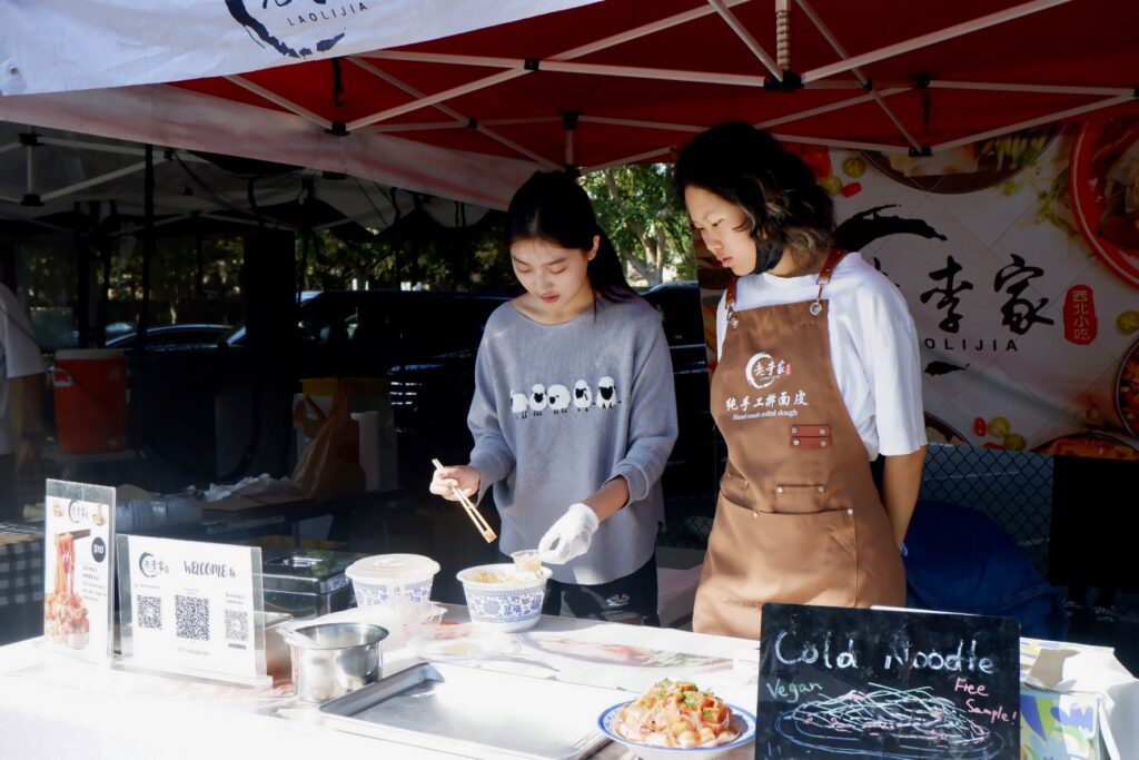 Farmers Markets 3 two young asian girls under a tent with one girl in grey sweater with sheep design holding chopsticks and the other girl in brown apron looking at what the other girl is doing