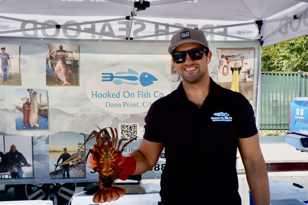 Farmers Markets 4 man in black polo shirt, grey cap and sunglasses smiling while holding a live lobster with a backdrop that shows hooked on fish co dana point, ca
