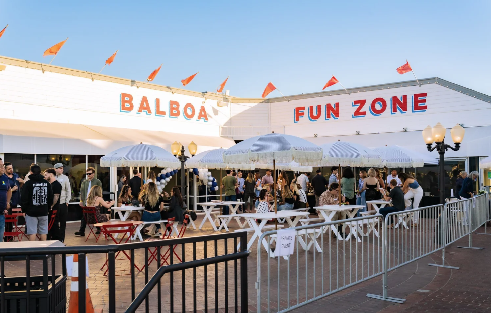 an enclosed square with tables and benches and people standing and sitting with Balboa Fun Zone painted on the wall with orange flags on the roof