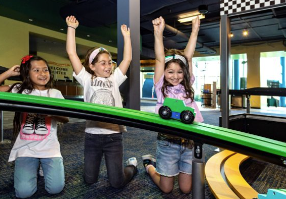 three girls playing on a toy track with two girls with their hands up watching a green and blue car traveling through the track