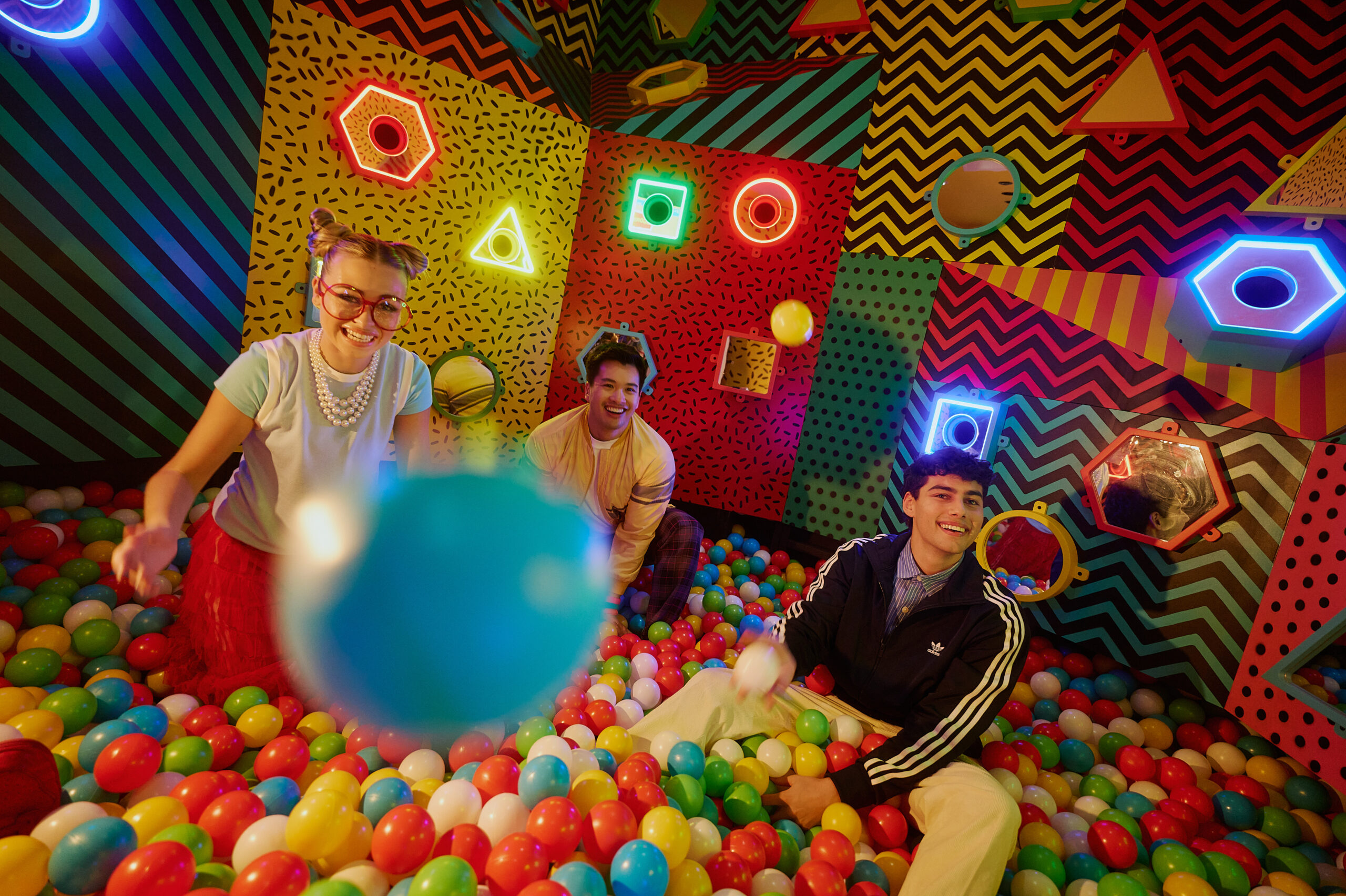 Colorful room with neon light shapes on the wall with colorful plastic balls to play into like a pool with two males and one female playing