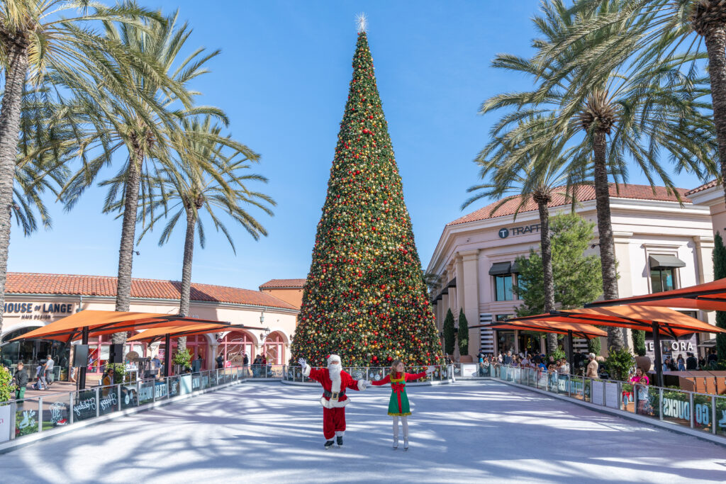 an outdoor ice skating rink surrounded by palm trees during daytime with santa clause and an elf on the ice, with a big christmas tree in the background