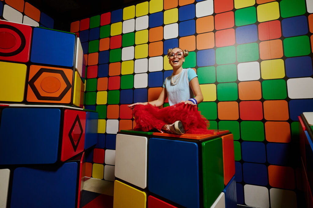 a woman in white shirt and red pants cross sitting on top of a big rubik's cube in a colorful playroom that resembles a rubik's cube