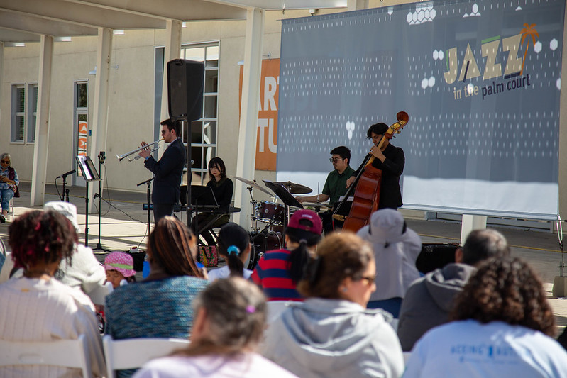a group of 4 musicians playing jazz in front of a crowd with a banner saying jazz in the palm court at the back