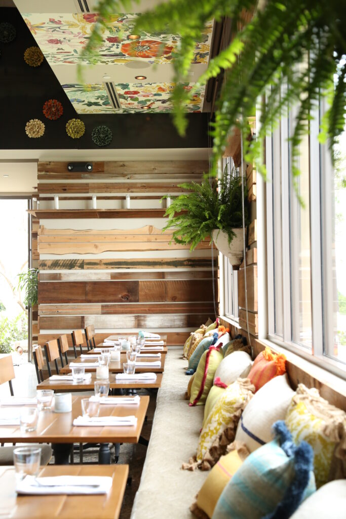 Discover Irvine’s Culinary Scene: From Breakfast to Dessert 3 a restaurant interior with an elongated connected seating area with colorful pillows against a glass window, with a hanging plant as decor