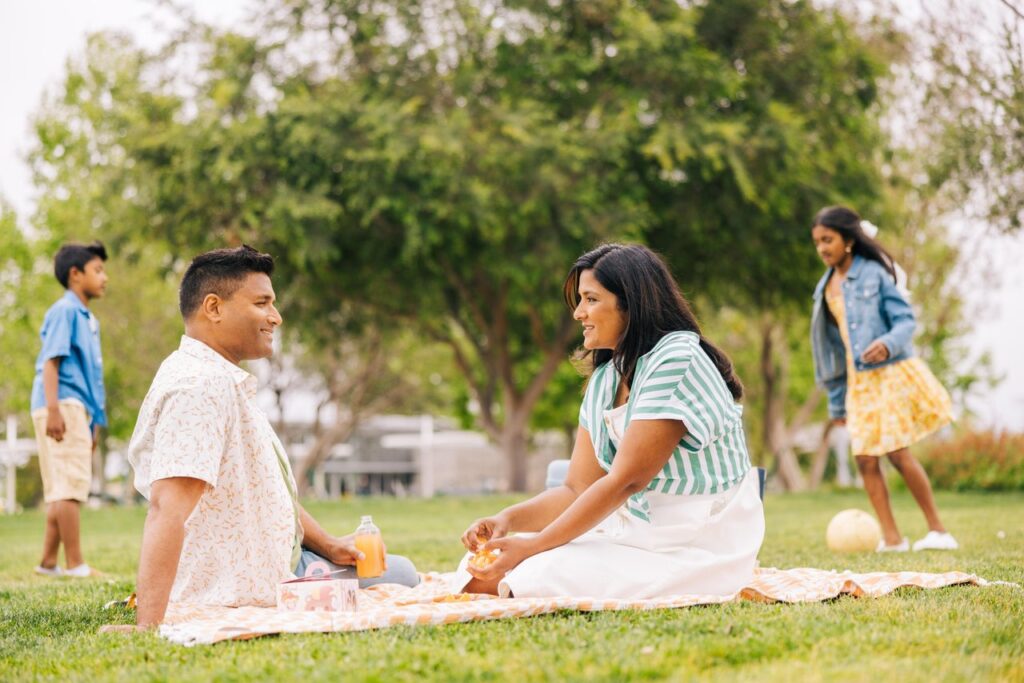 man and woman sitting on a blanket in a park having a picnic with son and daughter playing in the background at great park
