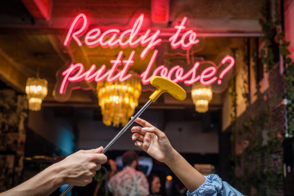 Neon sign of Ready to Putt Loose with chandeliers in the background with two hands holding a putt
