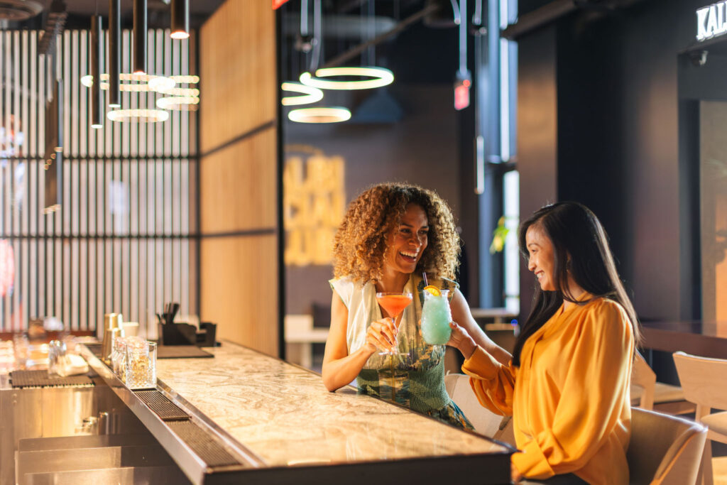 two women sitting at a restaurant bar toasting cocktails and smiling