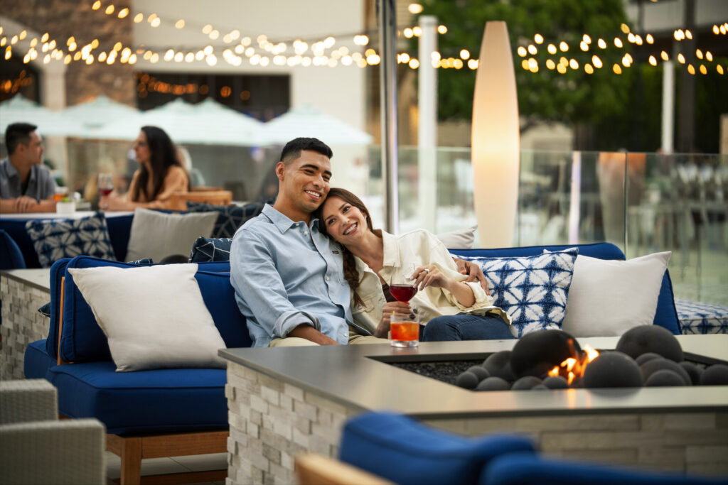 man and woman sitting on a couch with a firepit and night lights, with the omwn's head on the man's shoulder