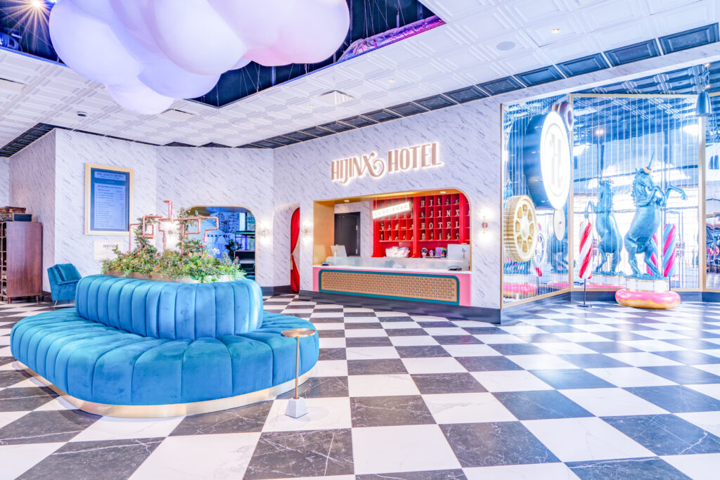 a colorful lobby with a front desk andhijinx hotel sign with blue lounge in the middle of the room and a blue unicorn statue on the side, a very whimsical space