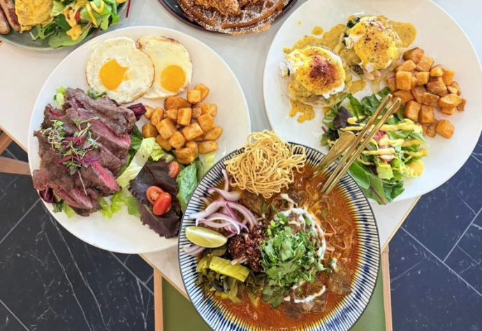 a table of food with two plates and one bowl, one plate with eggs, steak and potatoes, the other plate an eggs benedict, vegetables and potatoes, and a bowl of noodles