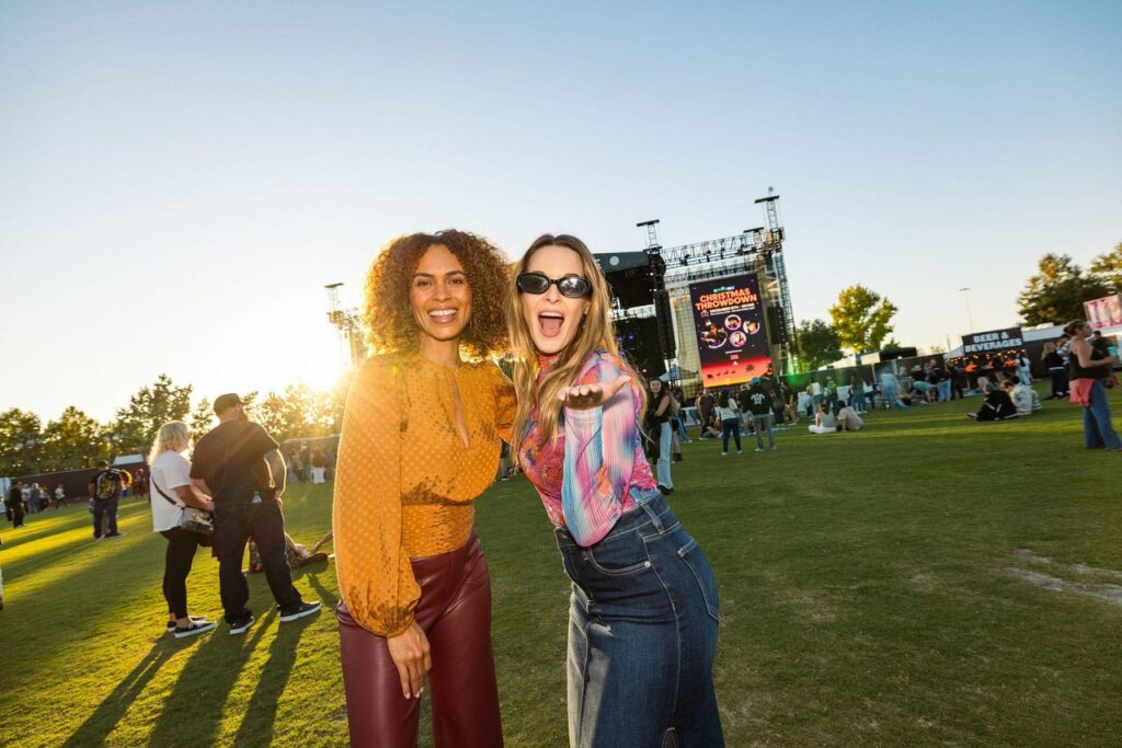 two ladies, one in orange with brown curly hair and the other with blonde hair and sunglasses in denim skirt and colored top at an outside event venue on a sunny day