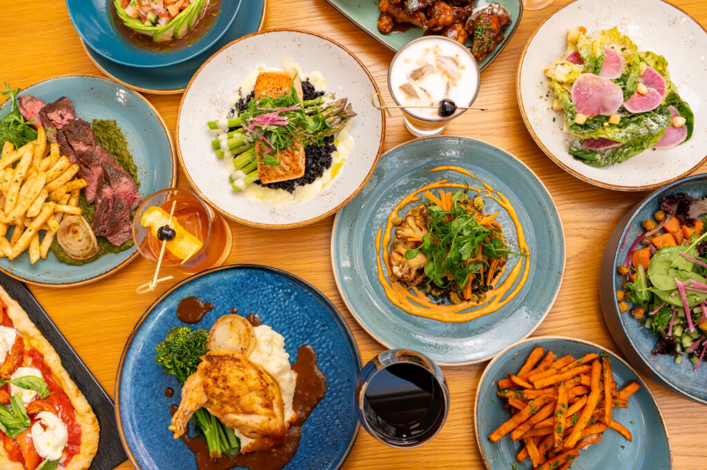 a table filled with plates with different food ranging from steak and fries, salmon and asparagus, salad, chicken, flatbread and cocktails