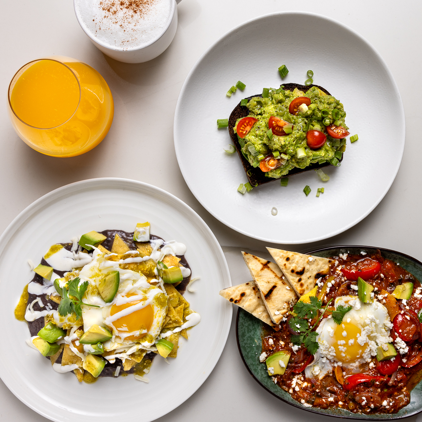 a table with orange juice, plate of avocado toast, chilaquiles, and another savory dish with pita and egg