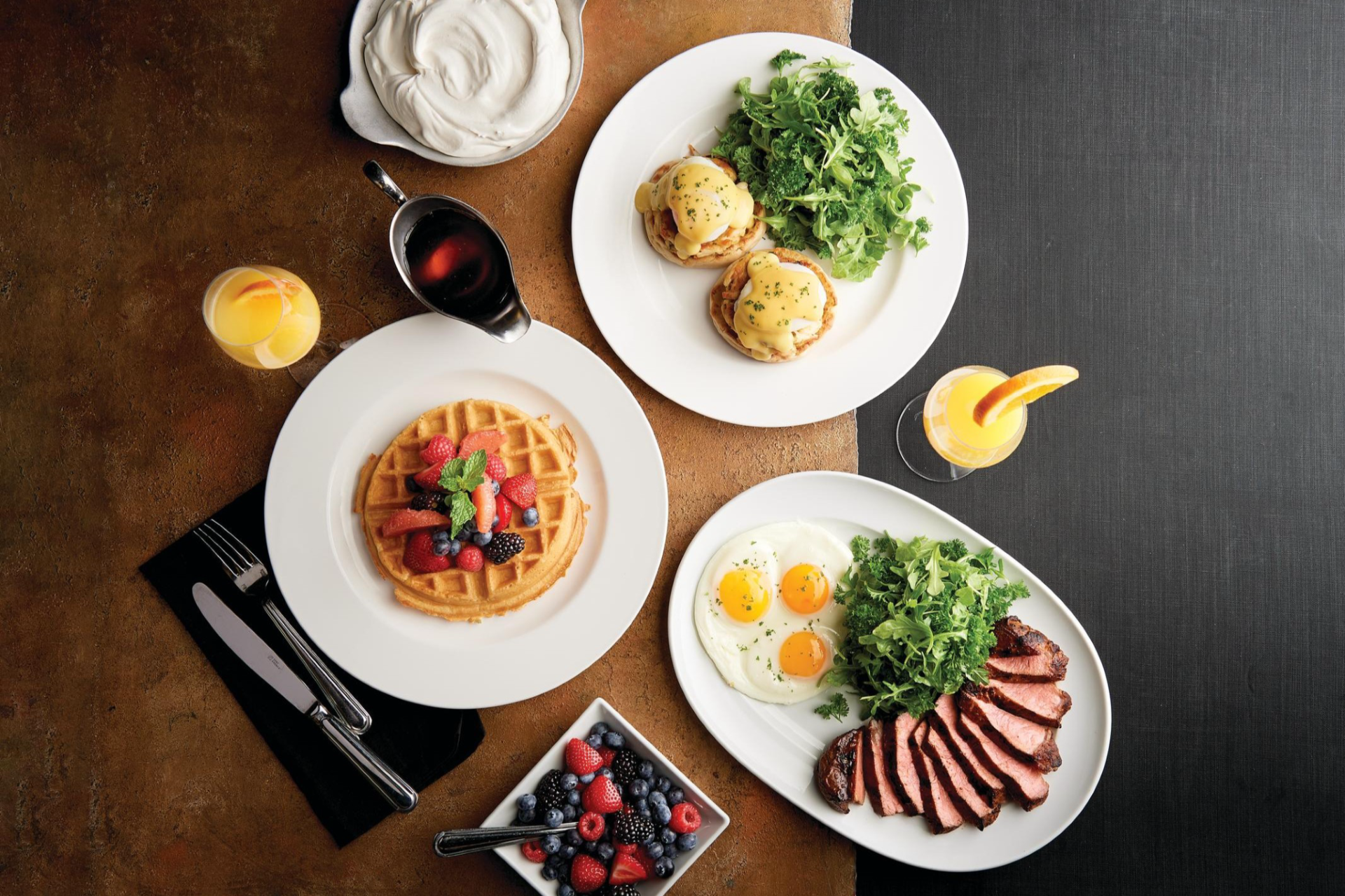 a table with a plate of eggs benedict and greens, a plate of waffle with strawberries, and a plate with eggs, greens, and steak with two glasses of orange juice