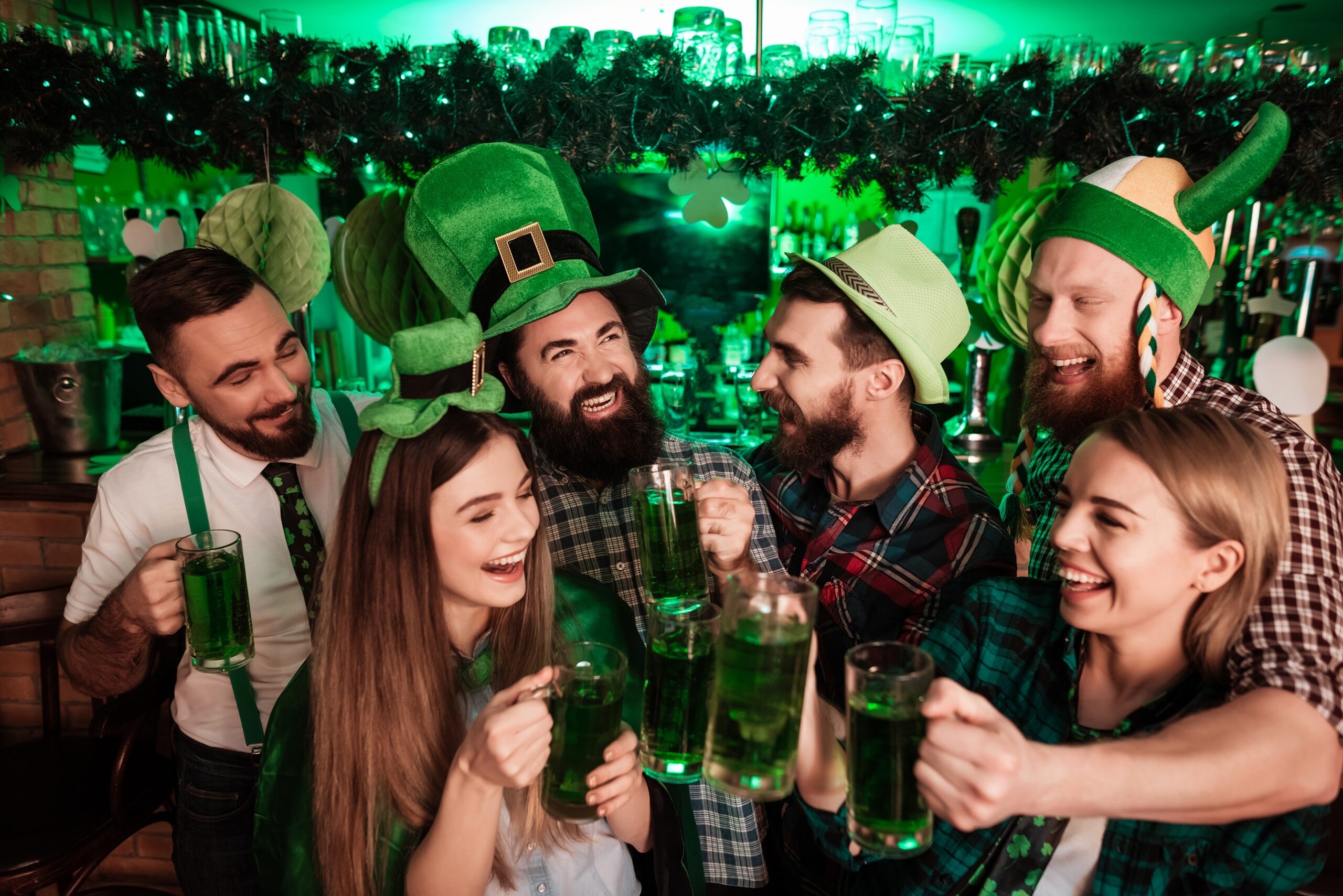 The company of young people celebrate St. Patrick's Day. They have fun at the bar. They are dressed in carnival headgear.