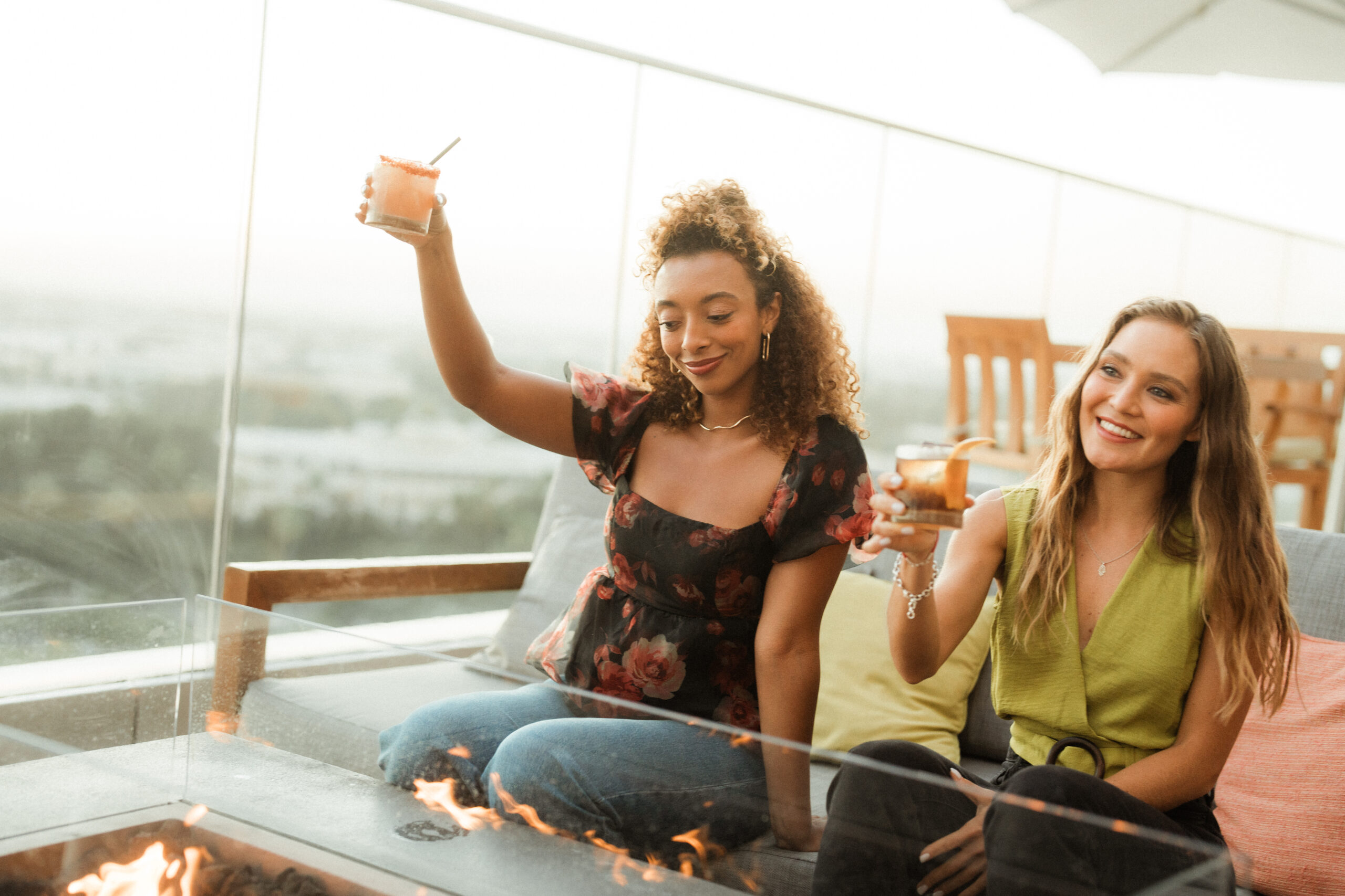 two girls sitting in front of a fireplace outdoors on a rooftop bar with cocktails in hand