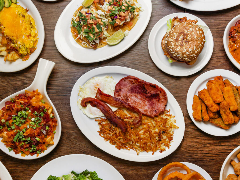 a table filled with plates of food ranging from a plate of tacos, burger, omelette with potatoes, fries, hash browns, bacon and egg