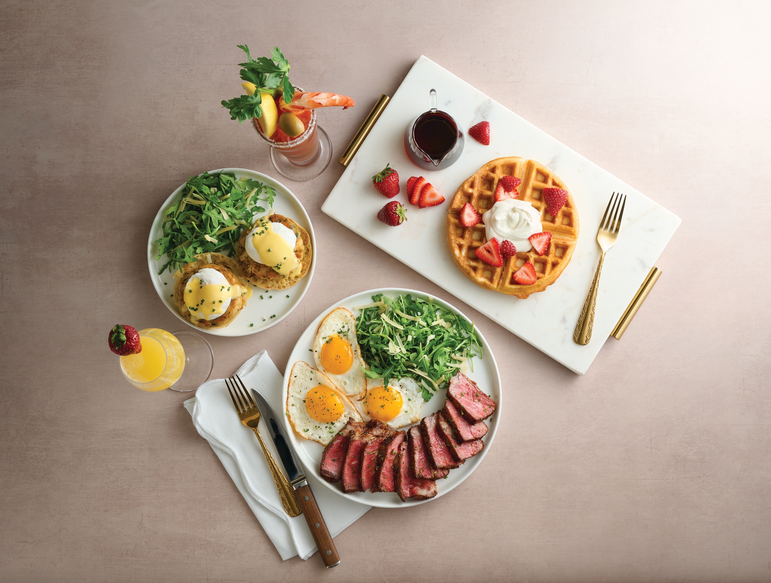 a table with a rectangular plate with waffle, a round plate with greens and eggs benedict, and another round plate with eggs, greens, and steak. two drinks are also on the table.