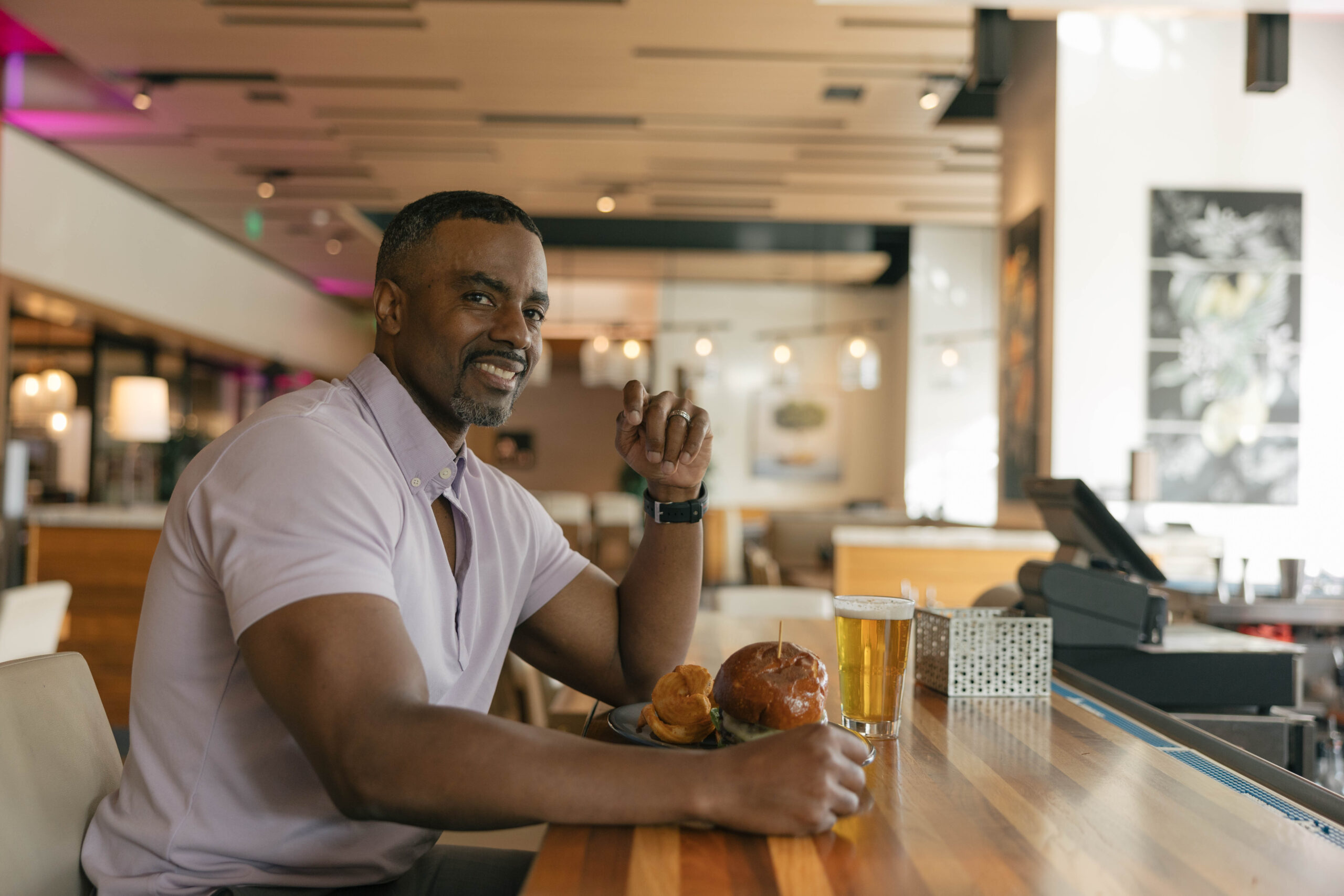 a muscular man smiling sitting a hotel bar with beer and burger in front of him