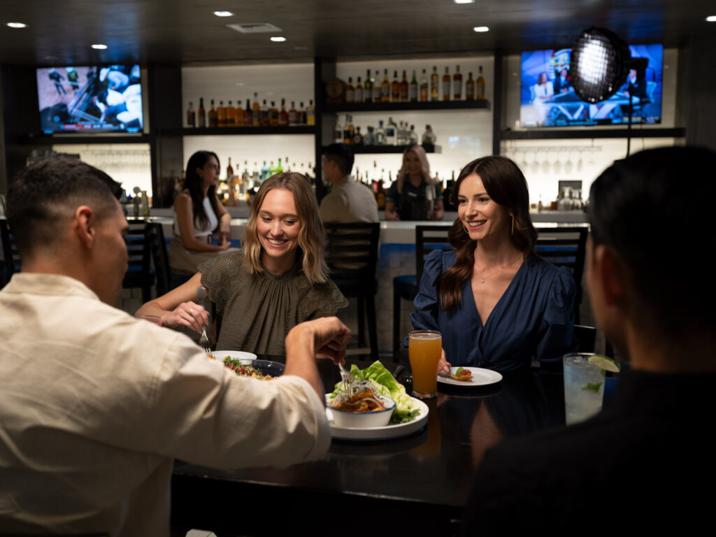 two females and two males with their backs sitting in a bar top with food and drinks in front, with the bar area behind them, having a night out