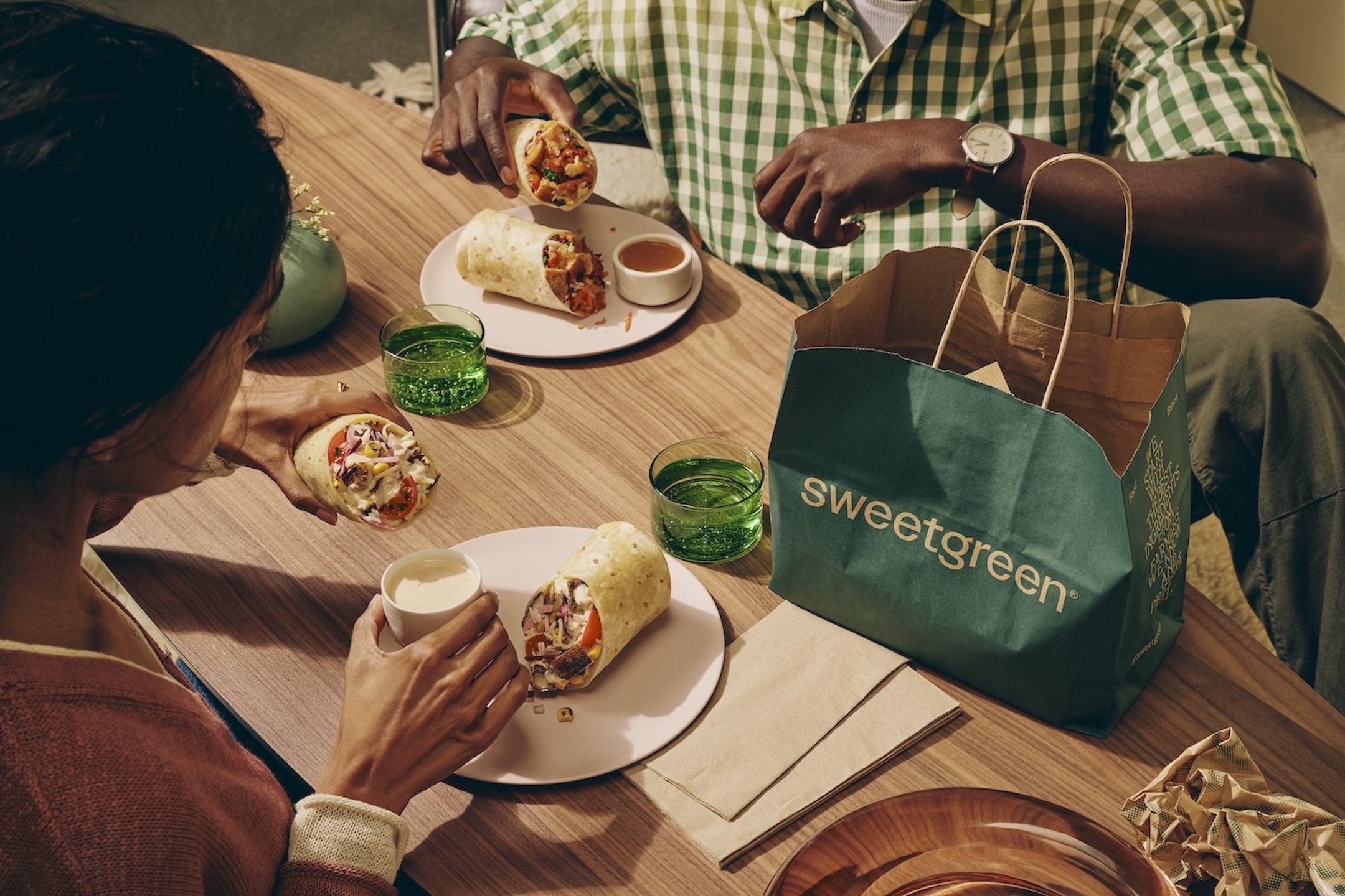 table with sweetgreen paperback, two people eating, showing their hands and two places with wraps