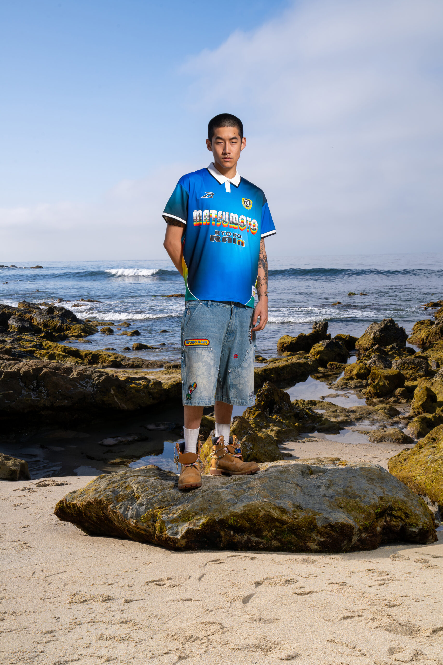 An Asian male in blue Matsumoto jersey and denim shorts standing on a rock at the beach