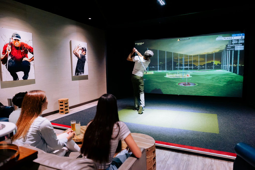 room with golf simulation with a male in full swing pose and two other people behind him sitting watching