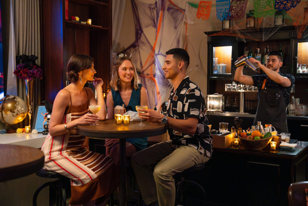two females and one male sitting in a high bar table with drinks at the table and the bartender mixing drinks behind them