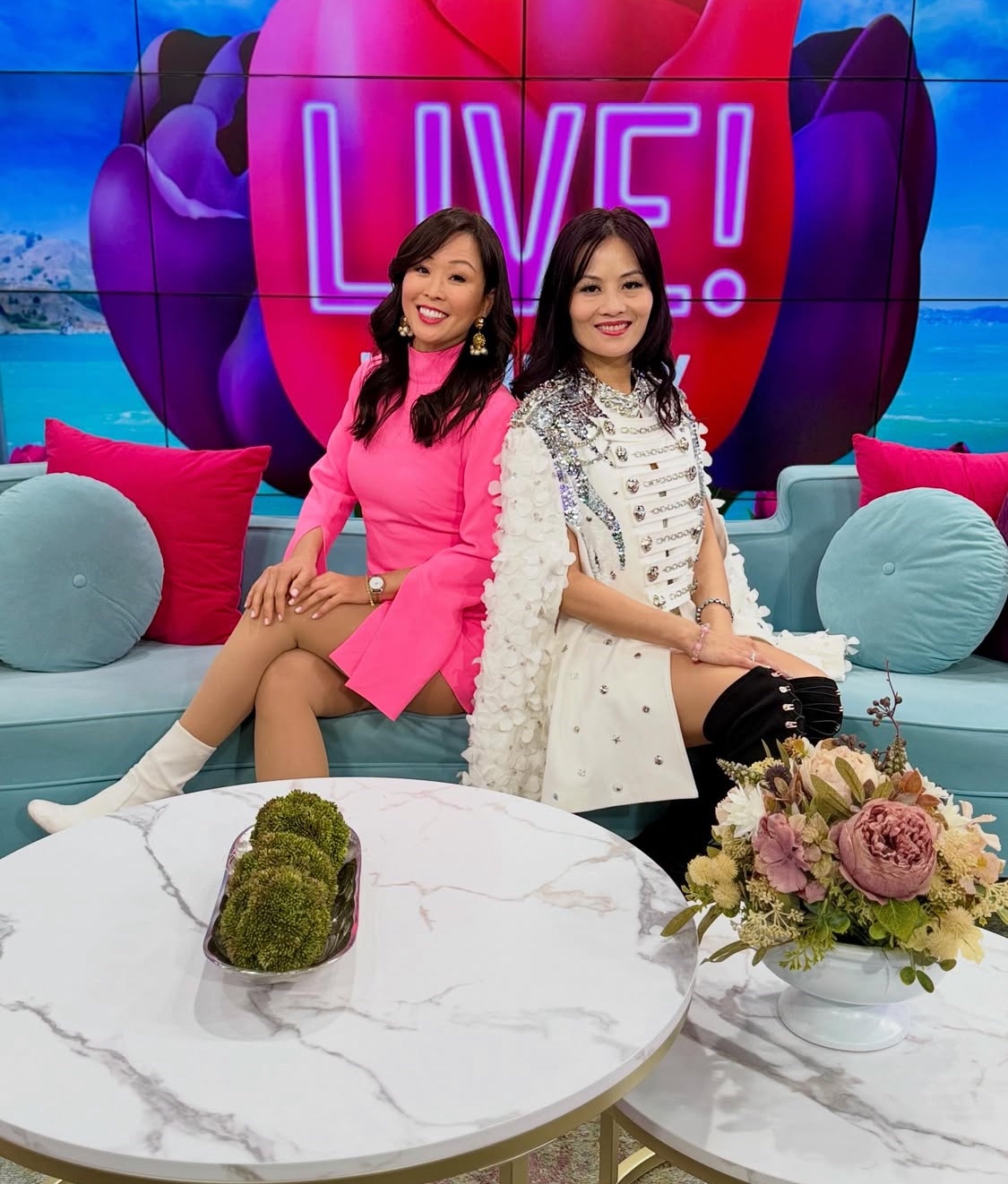 two Asian ladies one in pink dress and white boots and the other in white blazer and black boots sitting on a blue couch
