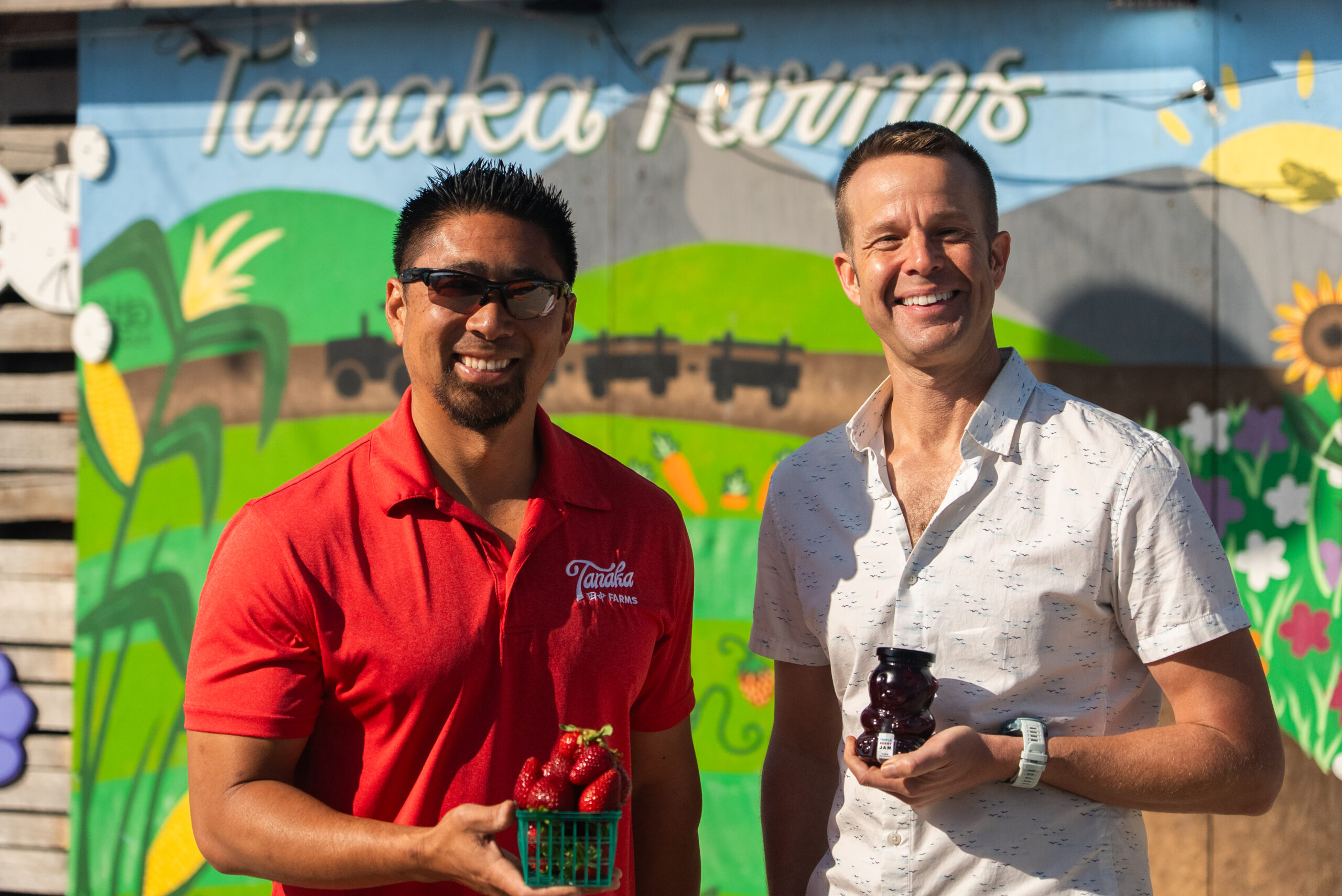 man in red polo shirt and sunglasses smiling holding a small basket of strawberries and a man in white polo shirt holding a bear shaped bottle of honey