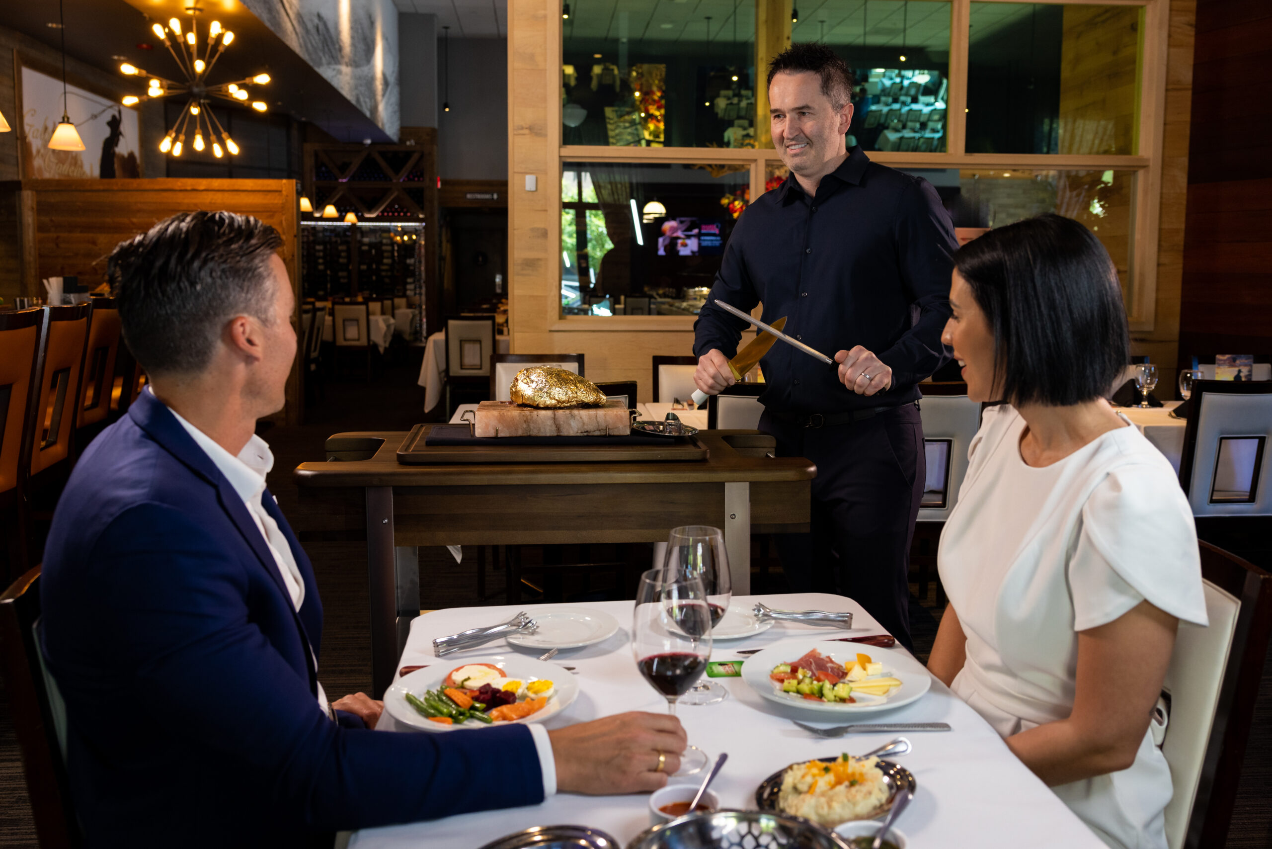 couple sitting on a table with a server preparing steak to serve