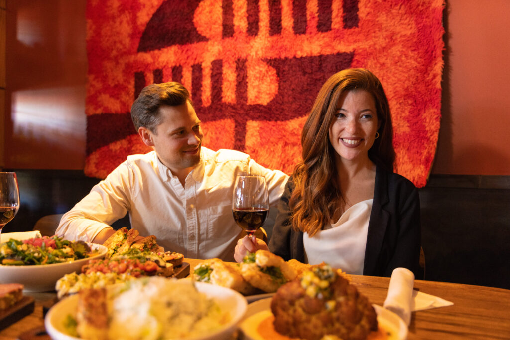 man and woman holding a glass of red wine in a restaurant with a table filled with italian food from bruschetta, to giant cauliflower at Postino Park Place