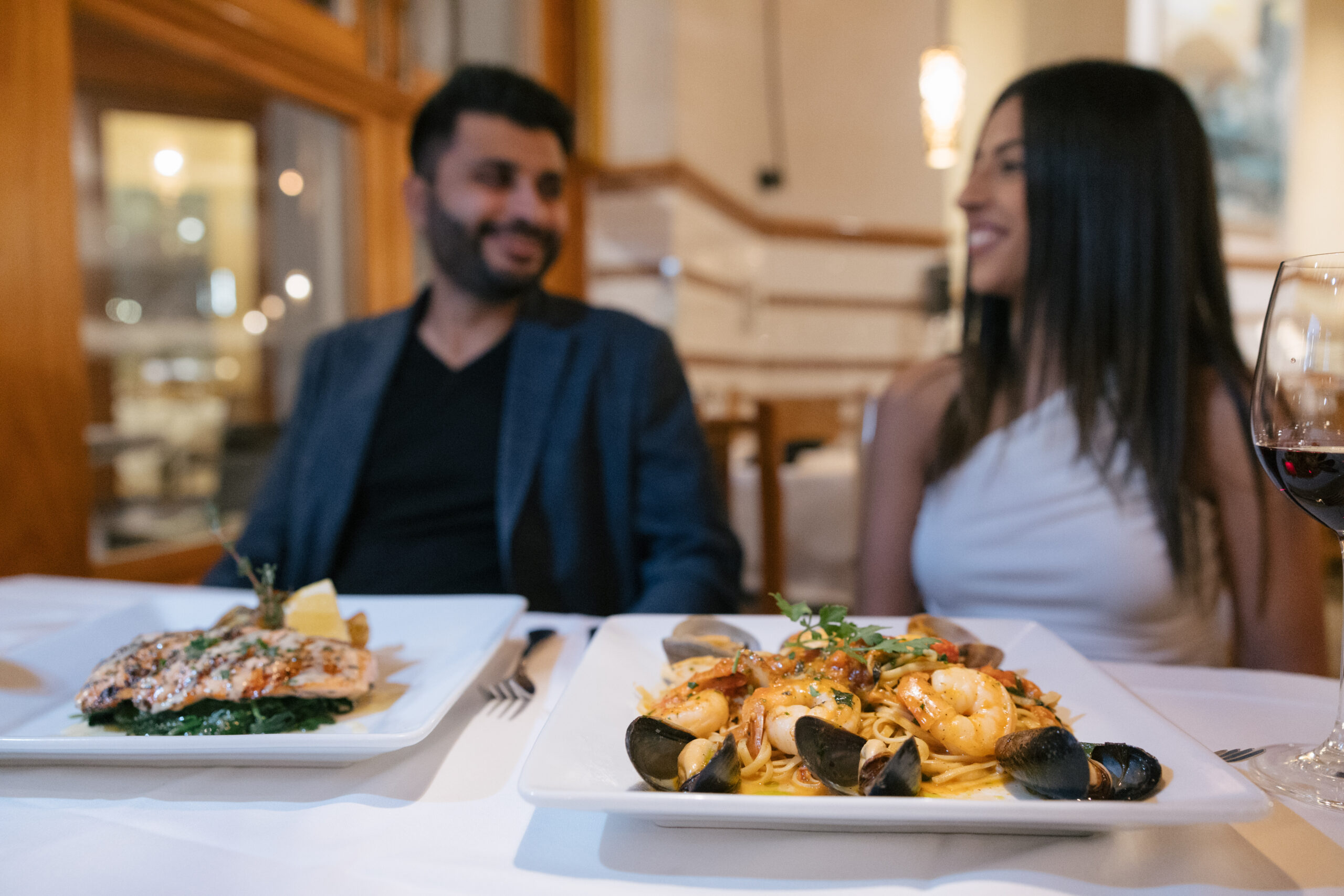 man in blue blazer and woman in white top sitting at a restaurant with a plate of salmon in front of the guy and seafood pasta in front of the woman at il fornaio restaurant